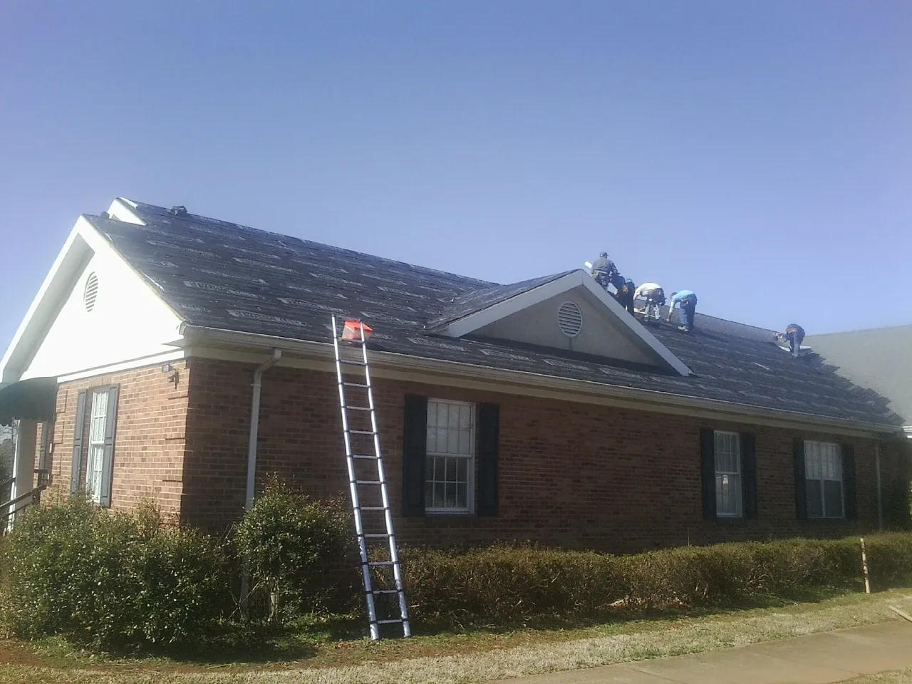 Crew tearing shingles off a brick ranch roof with a ladder set — Emergency Roof Repair work by our crew in Fort Dodge